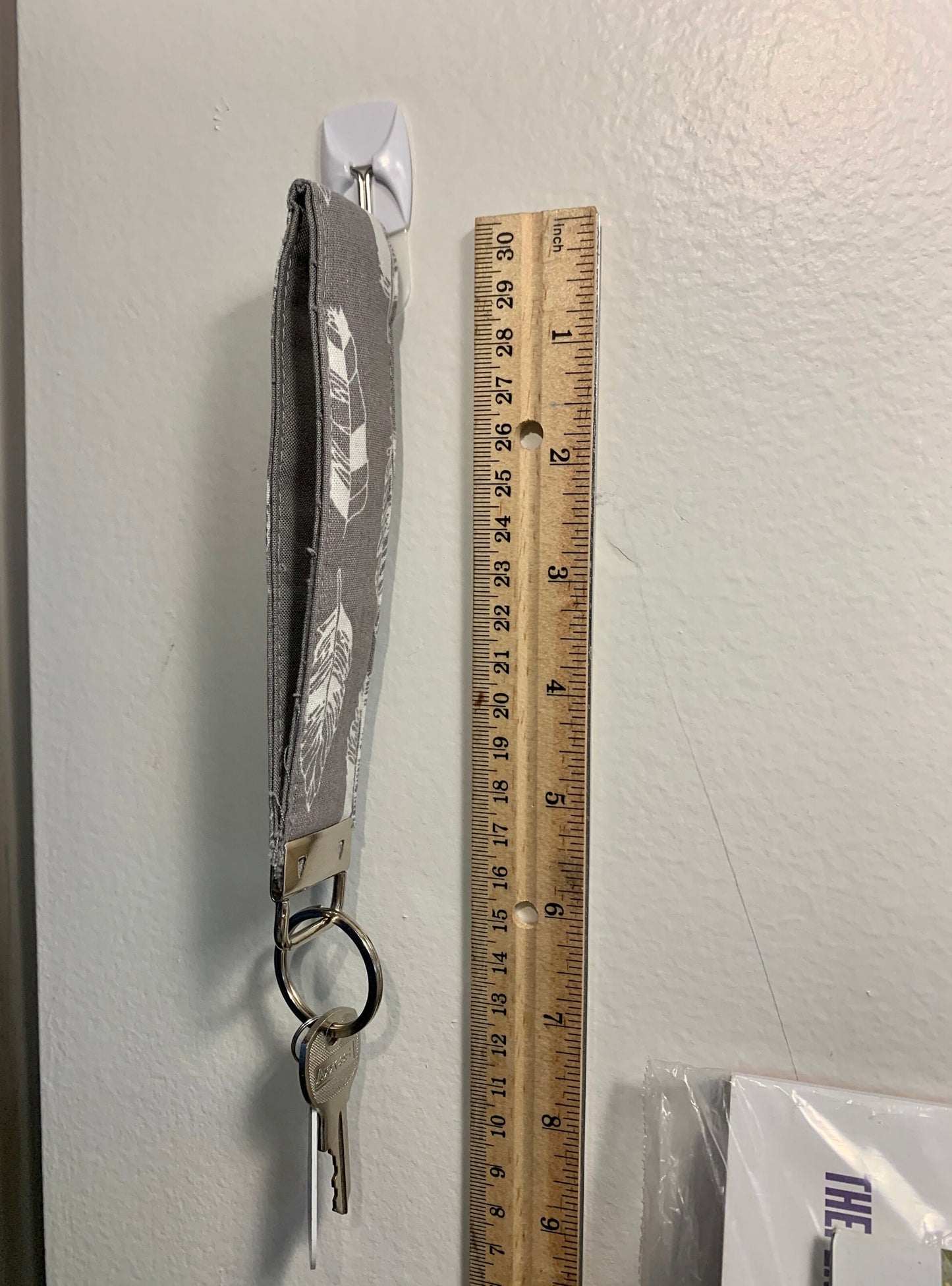 A grey keychain is shown hanging on a small command hook on the wall. A ruler is shown next to it, you can see that excluding the key ring it measures about 6 inches