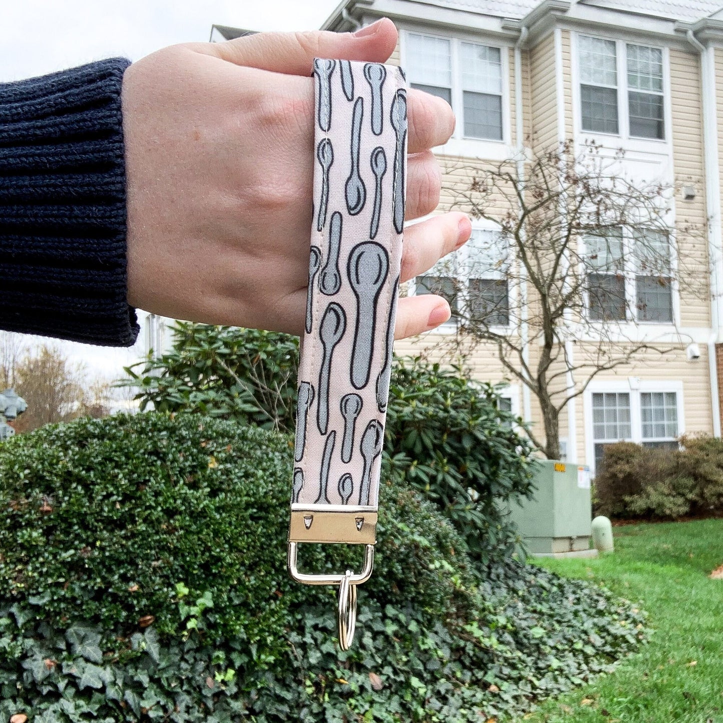 The cream spoons key fob is being shown held up in front of a building. It has silver spoons with black outlines, each spoon has one or two black brackets to show curvature. The pattern is on a cream background. The keychain is a loop of this fabric. It is held together at the bottom by a silver clasp with a bracket that hold a single key ring.
