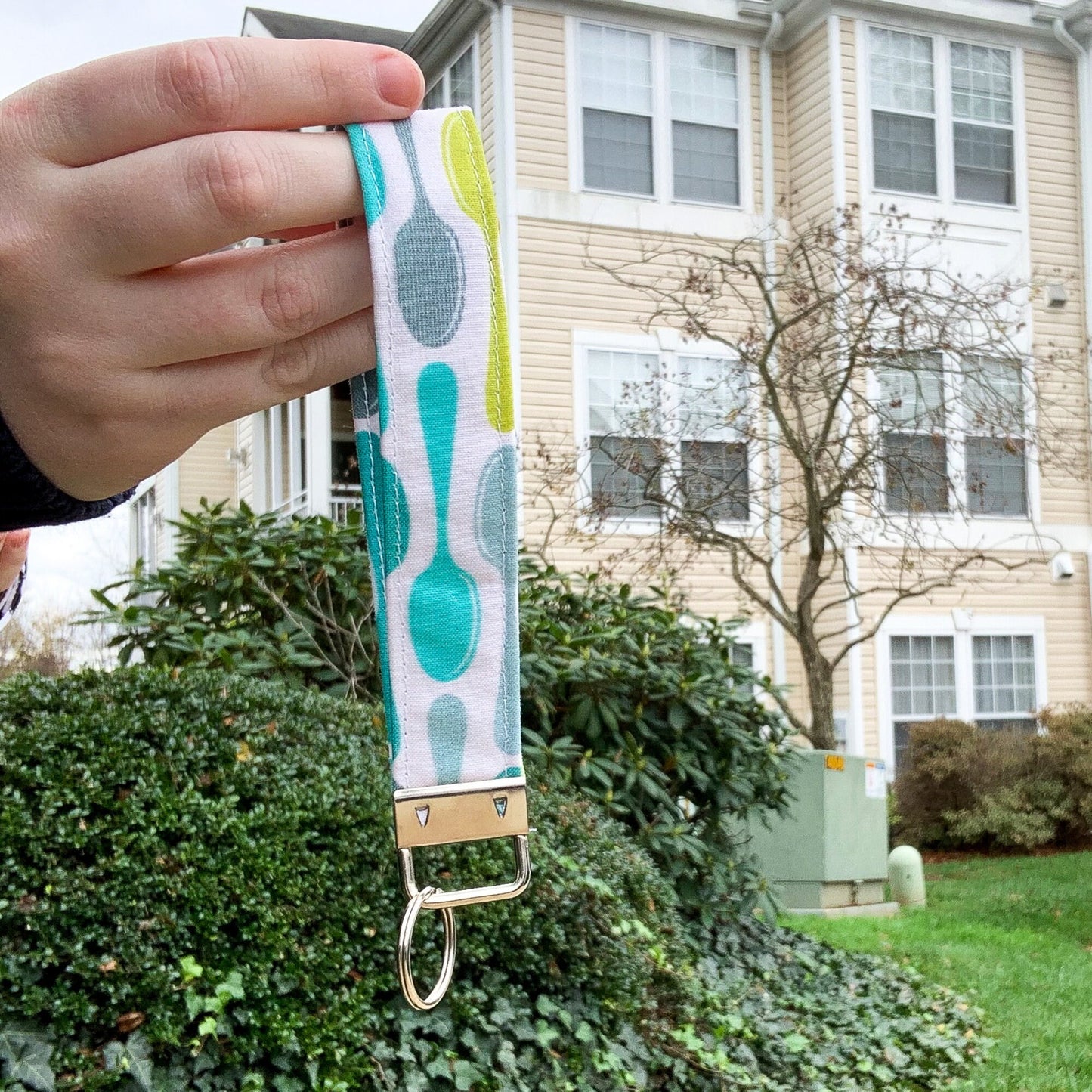 The blue spoons key fob is being shown held up in front of a building. It has lime green, blue grey, bright aquamarine, and teal colored spoons on a white background. The keychain is a loop of this fabric. It is held together at the bottom by a silver clasp with a bracket that hold a single key ring.