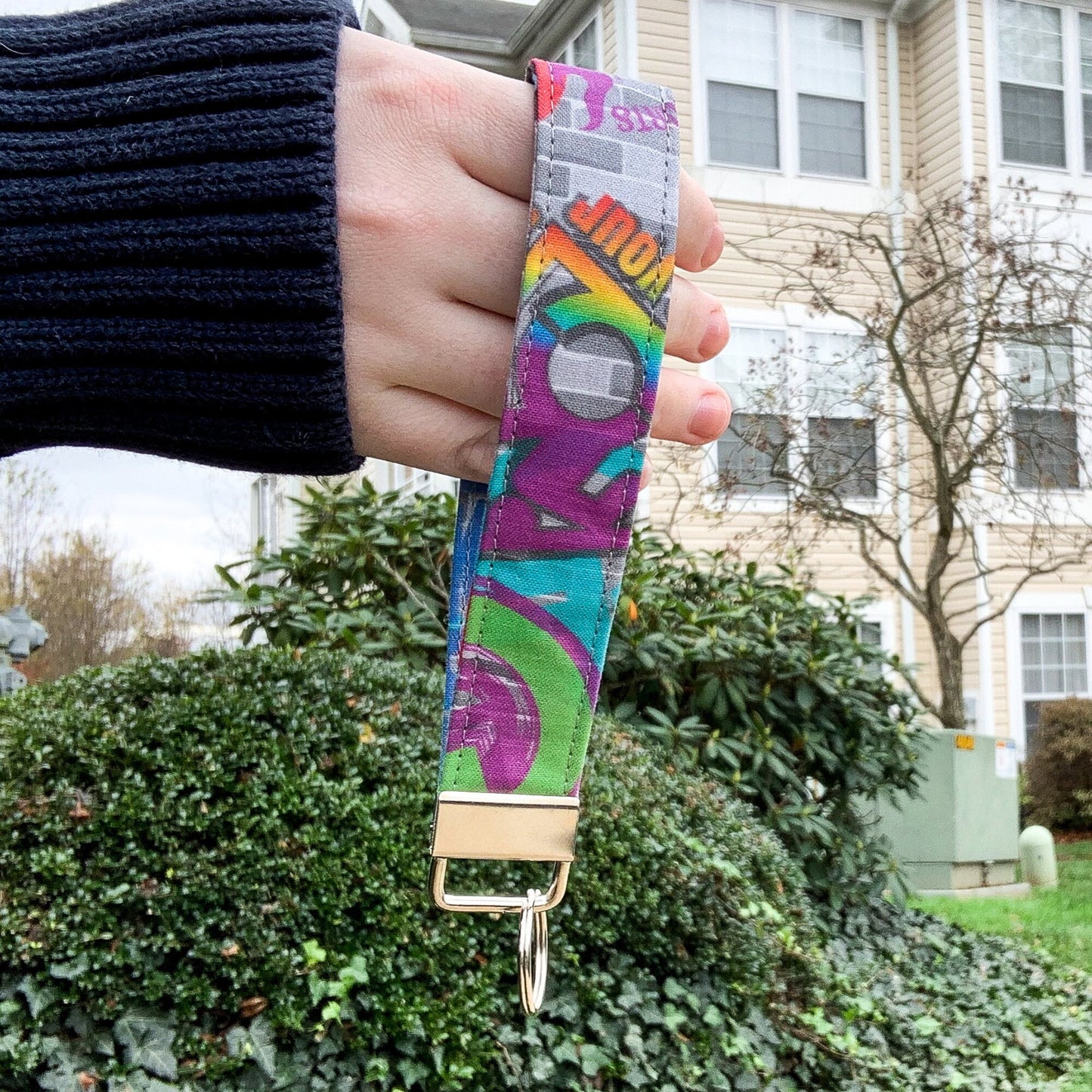 The adaptive graffiti key fob is being shown held up in front of a building. It has a rainbow disability symbol on this side and other text, it is on a grey brick background. The keychain is a loop of this fabric. It is held together at the bottom by a silver clasp with a bracket that hold a single key ring.