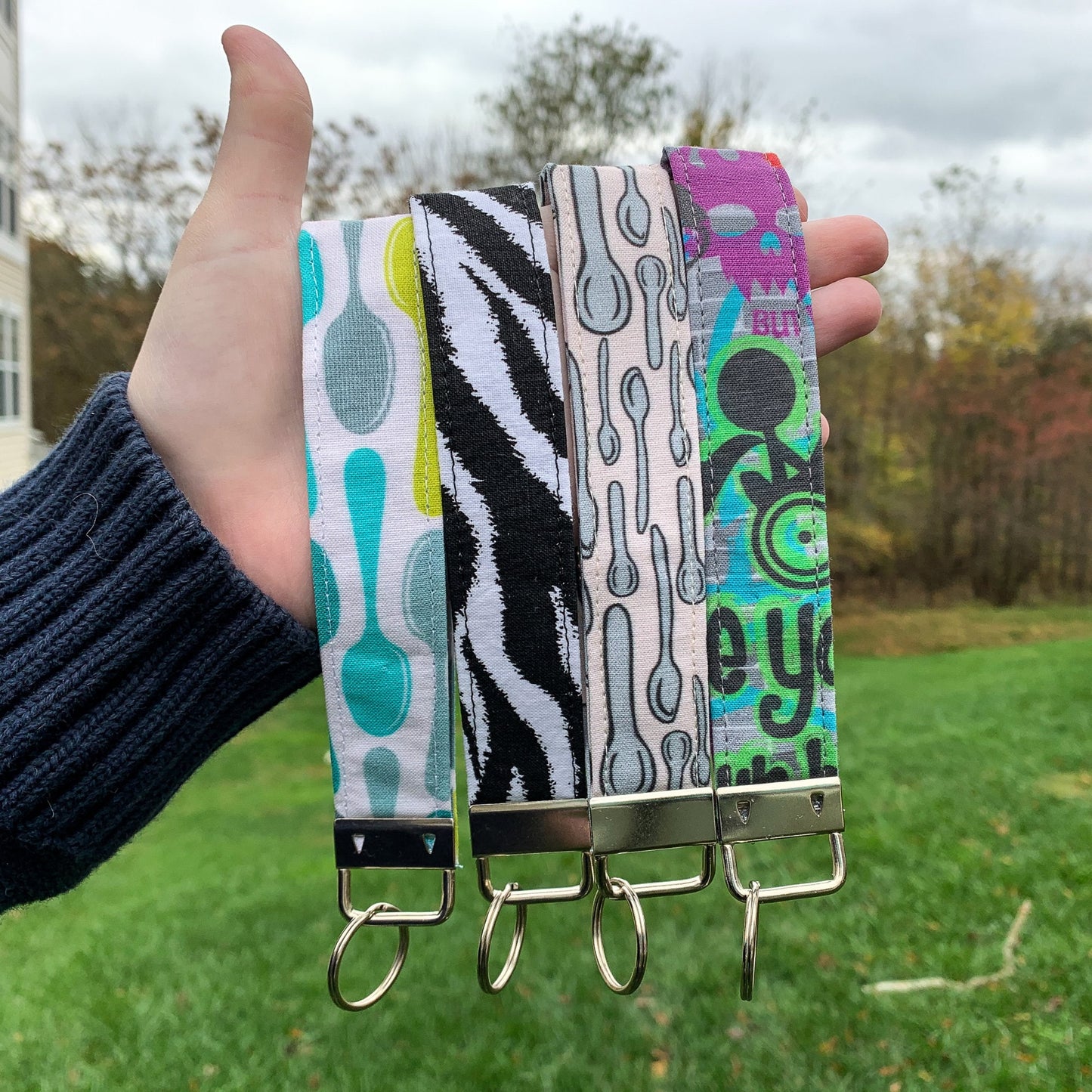4 keychain are being held up. From left to right the patterns are spoons in lime green blue teal and blue-grey on a white background, Zebra stripes, silver cartoonish spoons on a pinkish tan background, and graffiti with cartoonish drawings and lettering on a grey brick background. Each keychain has a metal clasp at the bottom with attached key ring.