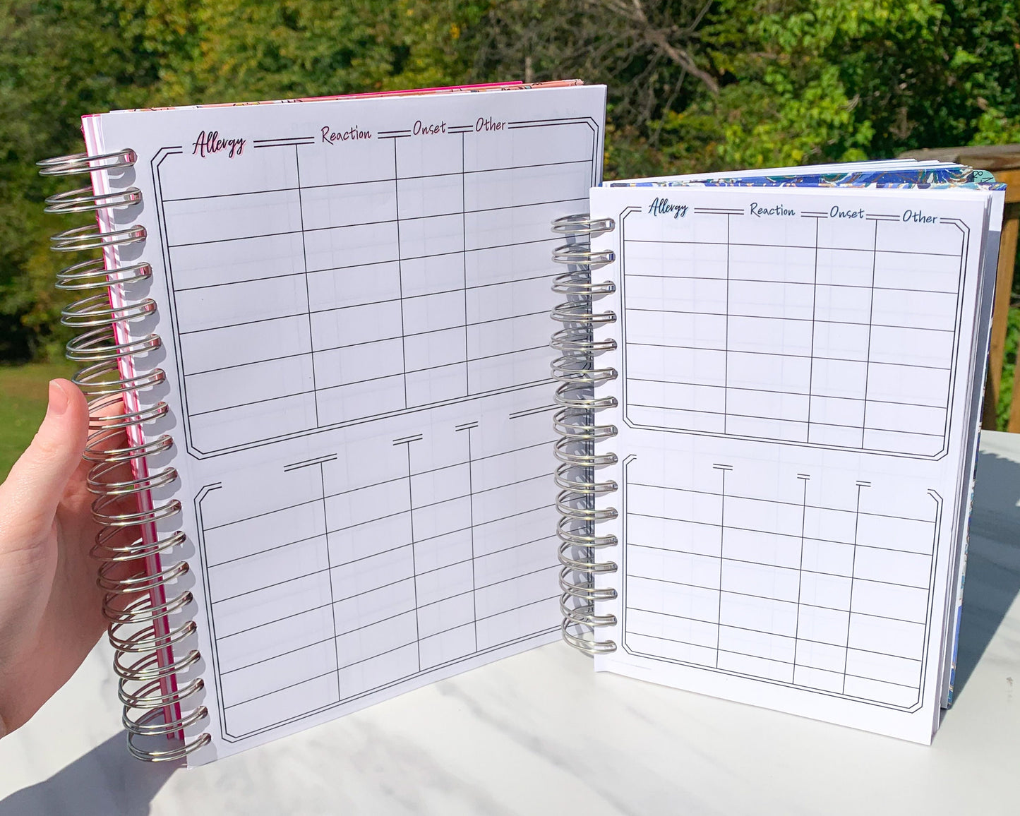 The larger pink and smaller blue books are being held up. They show the same 1 page. There are 2 boxes. The top one has columns labeled Allergy, reaction, onset, other. It has 7 rows. The bottom box has the same number of columns and rows but has blank slots to record whatever you want. The books have matching pink or blue shadows behind the text.