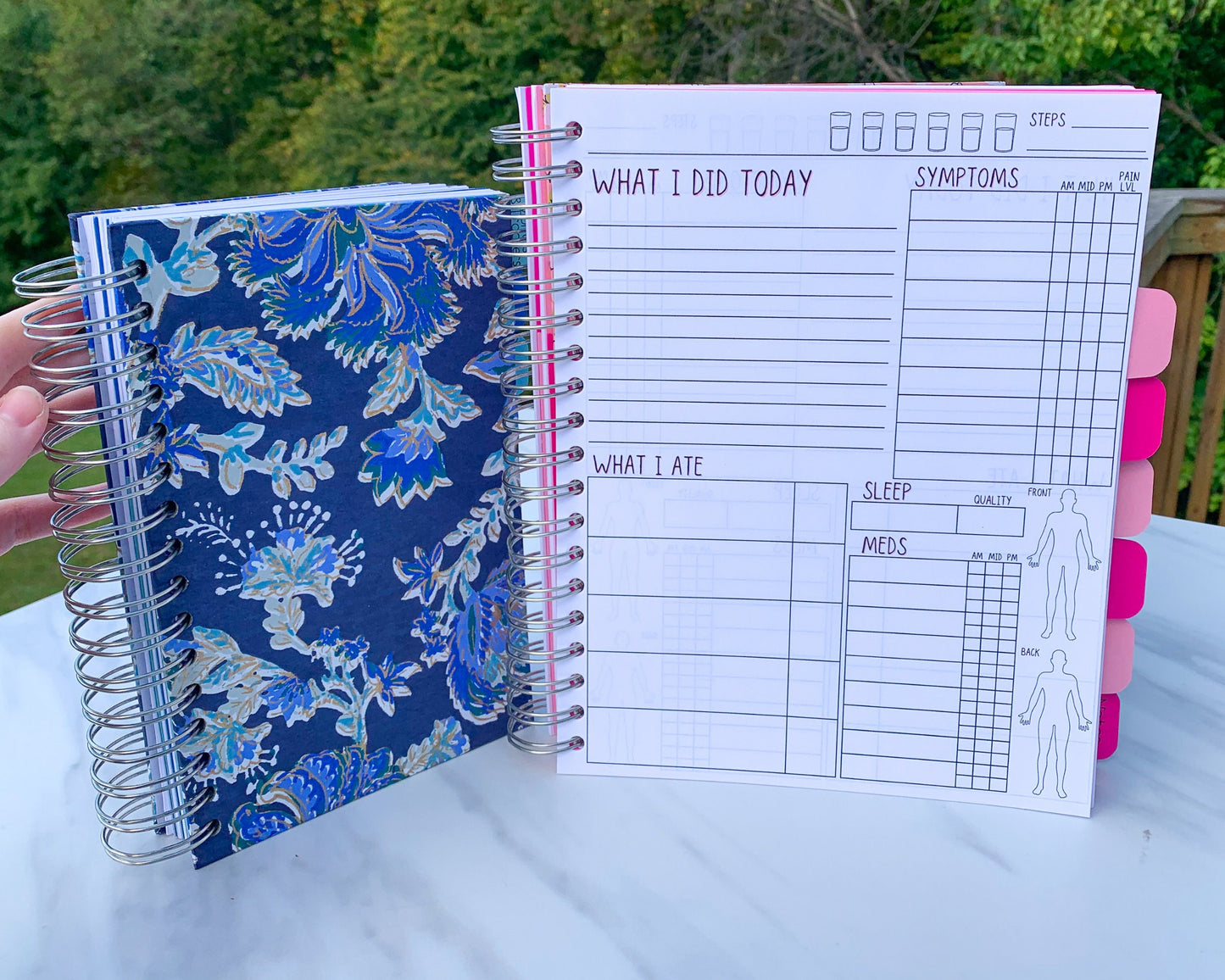 2 spiral bound books are standing up on a marble surface. The smaller one is closed and has a blue and gold floral cover. The other is open showing the interior pages. It has alternating light and dark pink tabs that are blank. The page has several sections and the titles have a pink shadow.