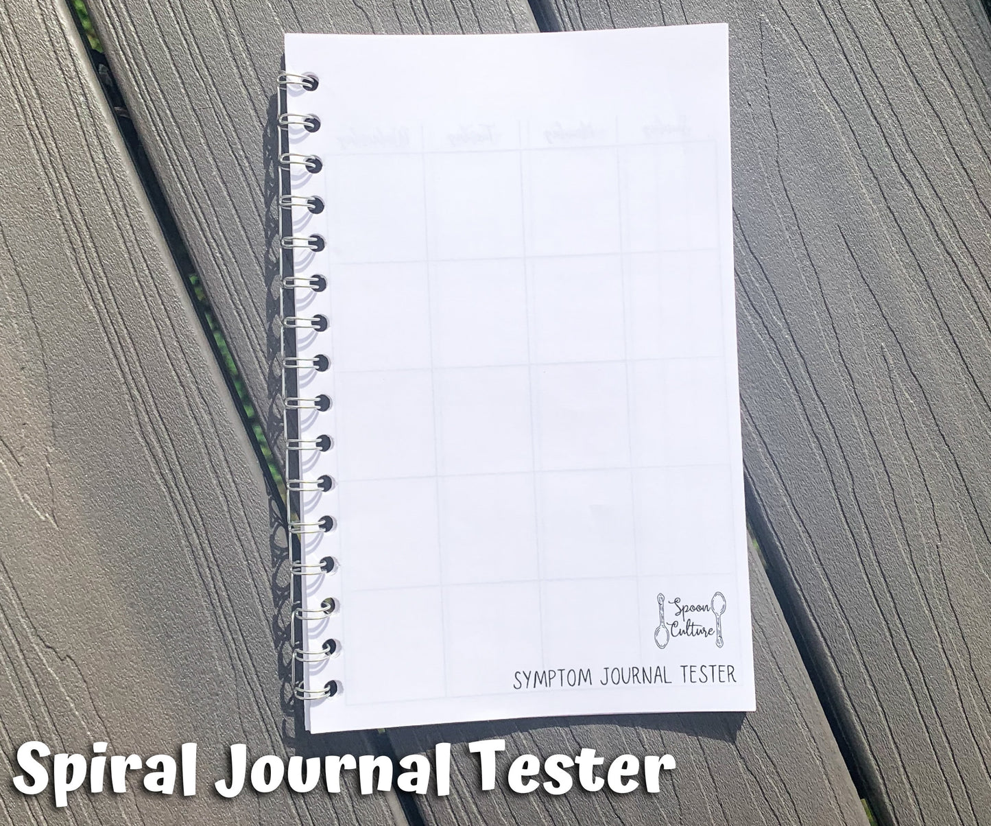 The spiral tester is shown on grey wood planks. It is closed and you can see the spoon culture logo on the front as well as the side of the coils. Text says spiral journal tester.