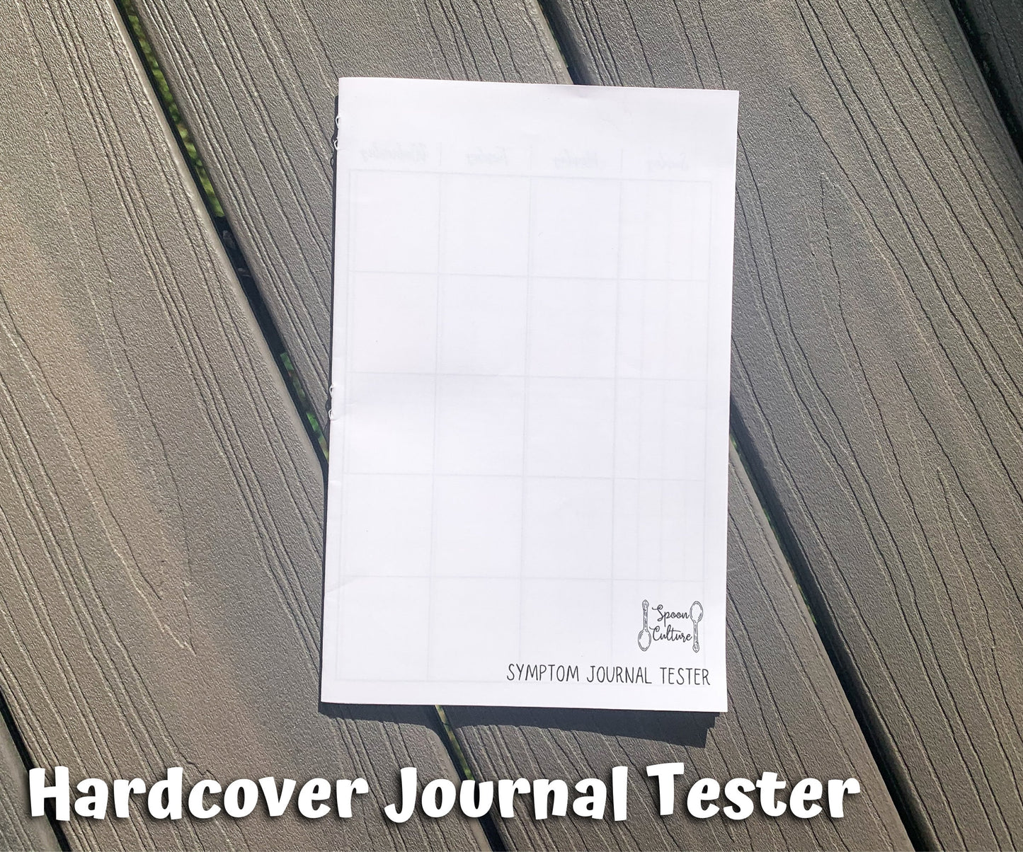 The hardcover tester is shown closed on grey wood planks. You can see the spoon culture logo on the bottom right and text that says symptom journal tester. On the left edge of the booklet you can see the back side of 3 staples. Text on the page says Hardcover Journal Tester.