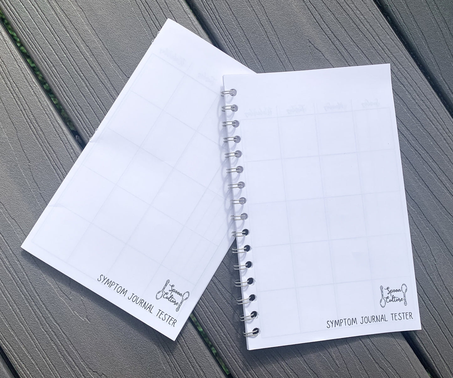 Both tester are closed on grey wood planks. Both have a logo in the bottom right corner that is two spoons in opposite directions with the words spoon culture in between, the words symptom journal tester are written below. The hardcover tester is on the left, it is flat folded pieces of paper. The spiral tester is on the right and you can see coils coming out of each hole punched.