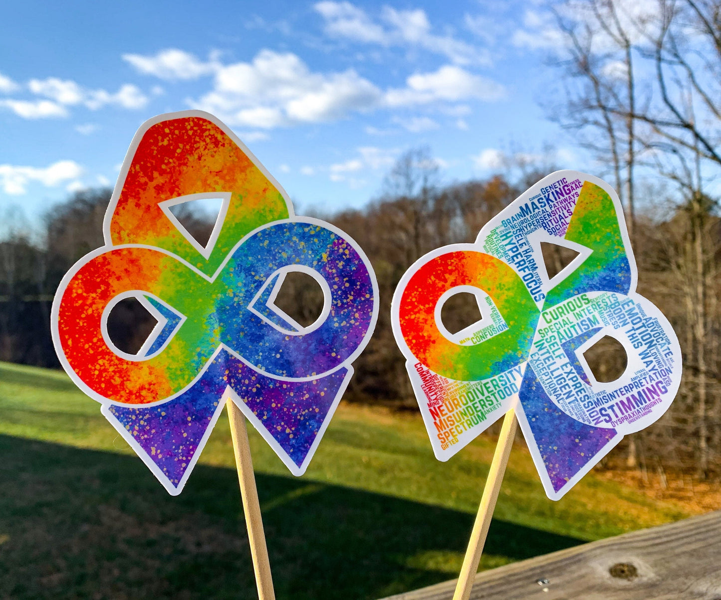 The word cloud and watercolor designs are shown on 2 sticks. Both ribbons feature an rainbow gradient awareness ribbon with a rainbow gradient infinity in the center. The color goes from red to purple left to right and there are gold flecks around the edges of the shapes. The word cloud design on the left is split in half with a word cloud on the left half of the ribbon and right half of the infinity.