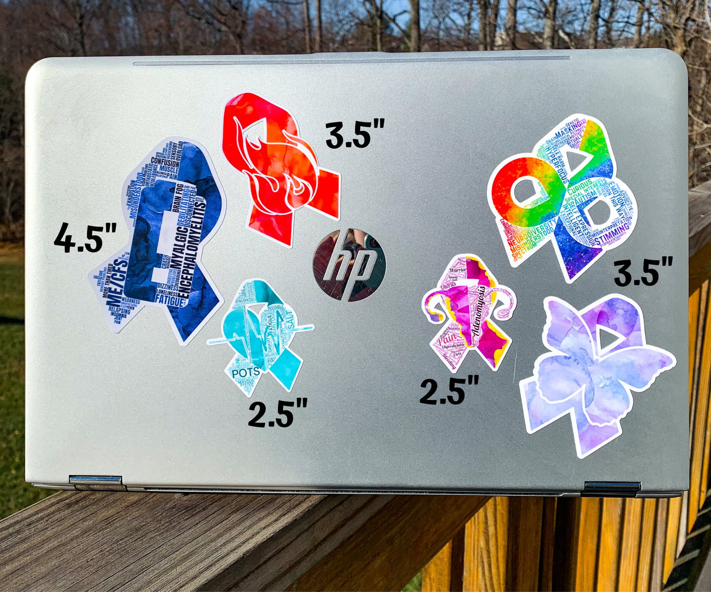 A silver laptop is shown with several stickers for size comparisons. There are 2 1/2 inch Adenomyosis and POTS stickers, 3 1/2 inch CRPS and Fibromyalgia stickers, and 4 1/2 inch Neurodiversity and ME/CFS Stickers
