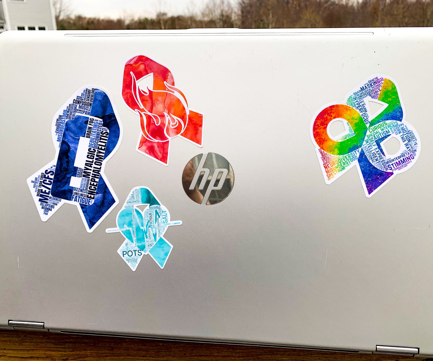 A silver laptop is shown with a 4 1/2 inch word cloud MECFS ribbon compared to a 3 1/2 inch watercolor CRPS and word cloud autism ribbon and a 2 1/2 inch word cloud POTS sticker.