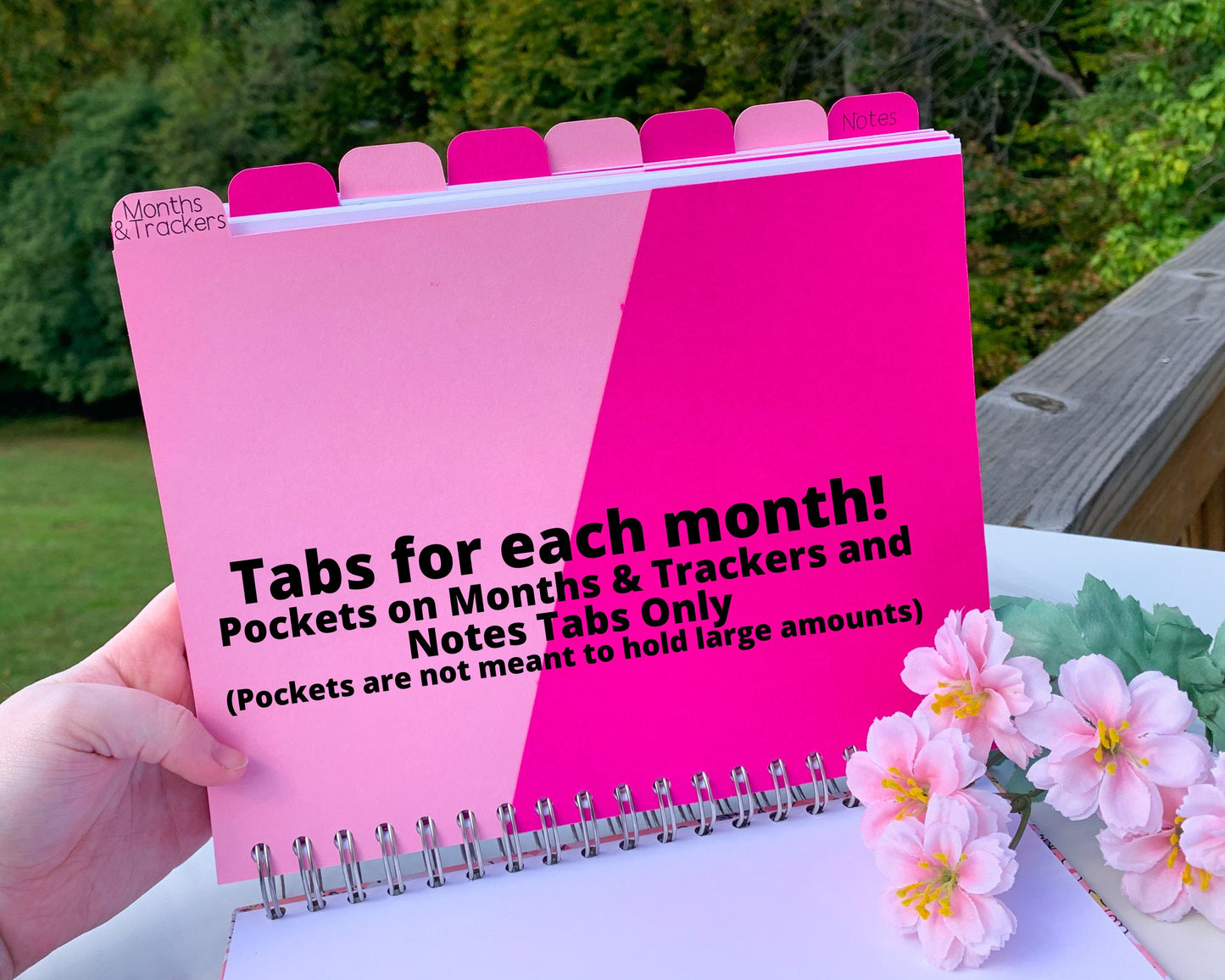 The book is being held up without the cover to show the tabs. The first tab is labeled Months and trackers, it is light pink with a darker hot pink angled pocket. The other tabs are blank and the last is labeled Notes. The tabs alternate between light blue and hot pink, with black writing. A note says Tabs for each month, pocket on months and tracker, and notes tabs only. Pockets are not meant to hold large amounts.
