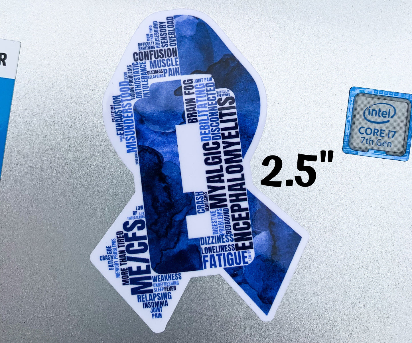A 2 1/2 inch MECFS ribbon is shown on a silver laptop. It is next to a sticker that says Intel Core i7 to show size comparison to the small stickers on a computer.