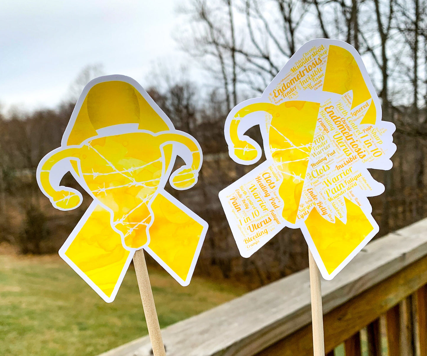 The word cloud and watercolor designs are shown on 2 sticks. Both ribbons are yellow with a shape in the center, the left watercolor one is a full uterus with barbed wire and the word cloud is half uterus and barbed wire and half sunflower. The word cloud design on the left is split in half with a word cloud on the left half of the ribbon and right sunflower half. The right watercolor ribbon is all color with a white border around the uterus for definition.