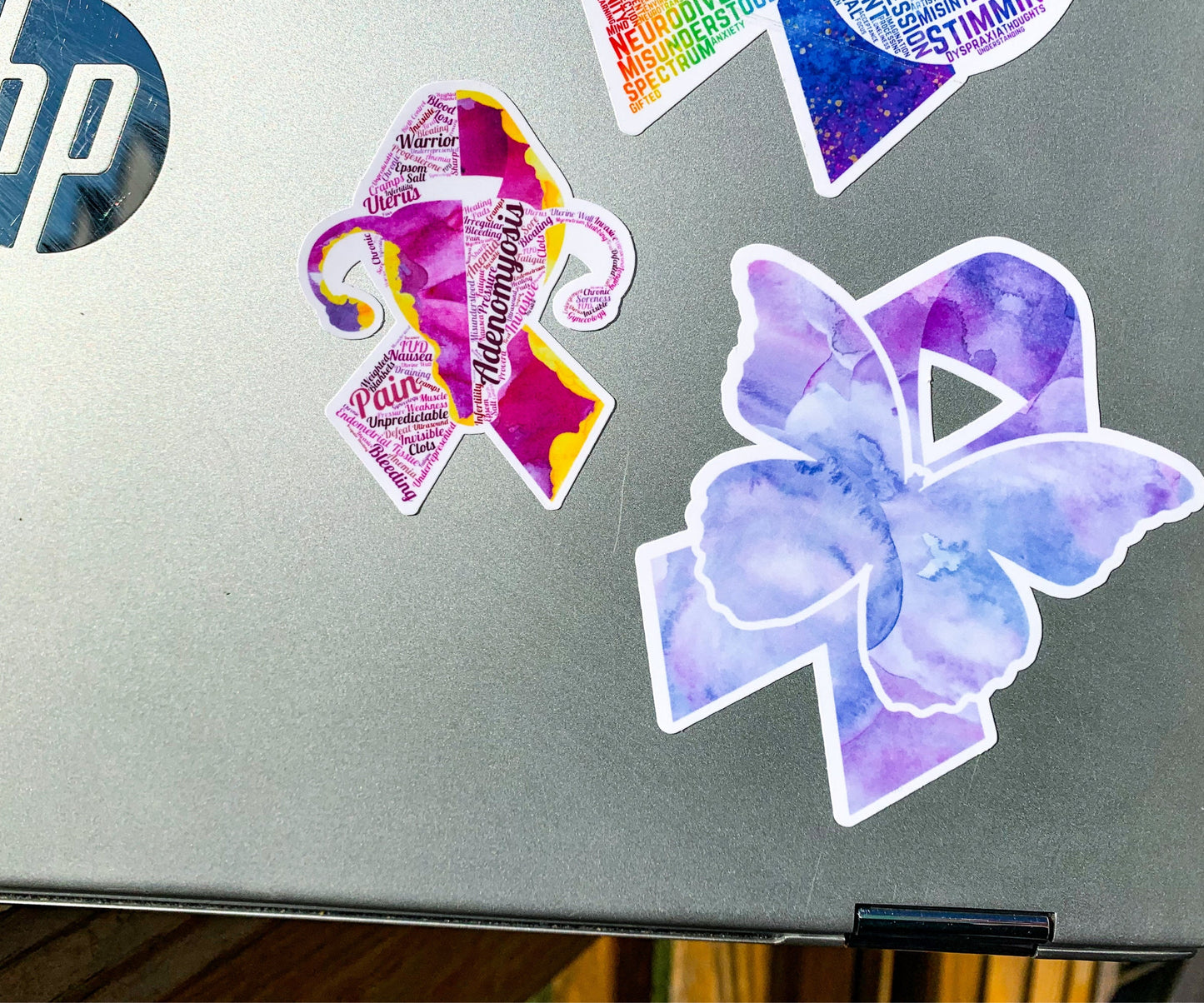 The Adenomyosis and Fibromyalgia ribbons are shown on a silver laptop. The Adenomyosis ribbon (left) is shown in size comparison to the larger Fibromyalgia (right) sticker.