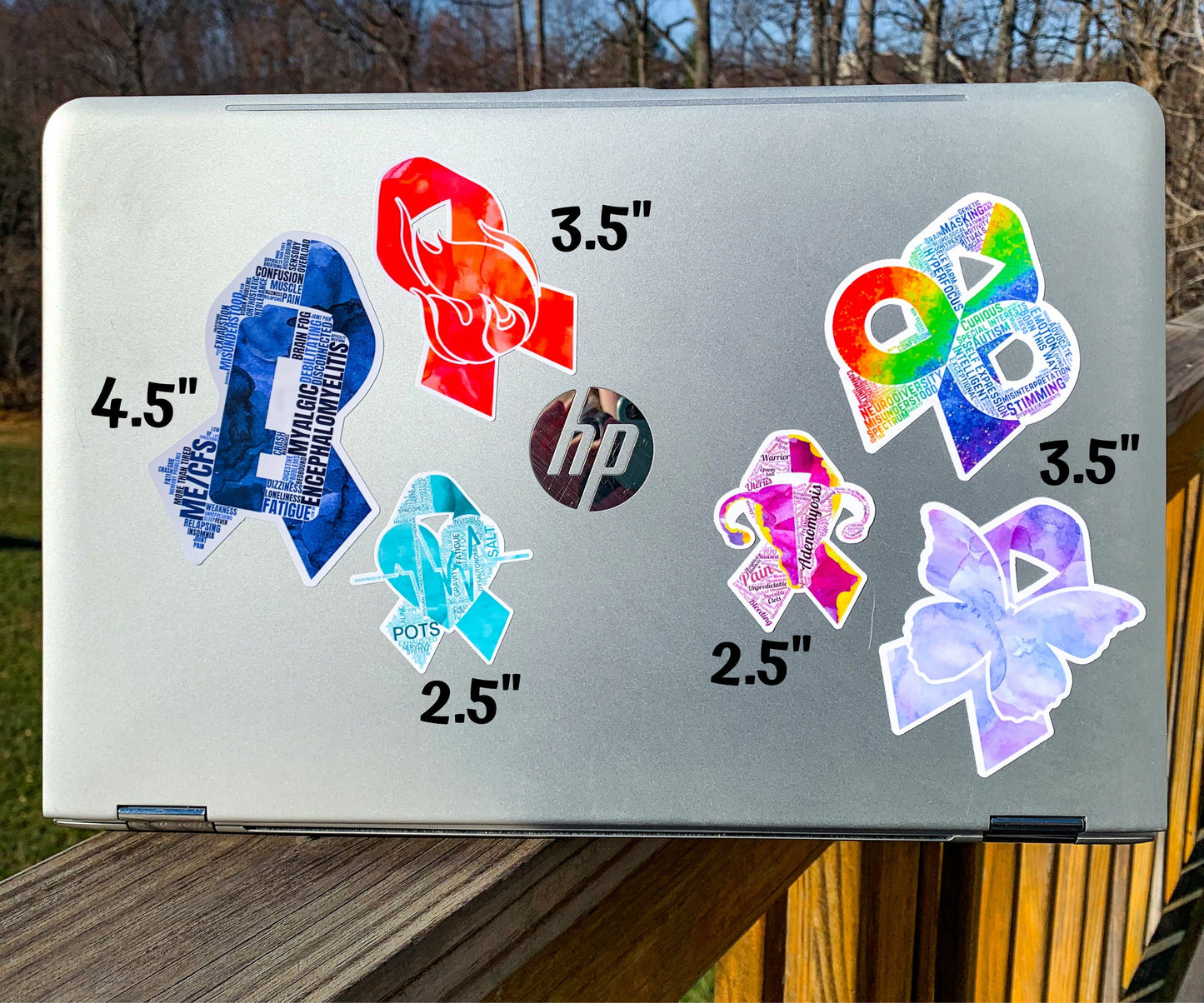 A silver laptop is shown with several stickers for size comparisons. There are 2 1/2 inch Adenomyosis and POTS stickers, 3 1/2 inch CRPS and Fibromyalgia stickers, and 4 1/2 inch Neurodiversity and ME/CFS Stickers