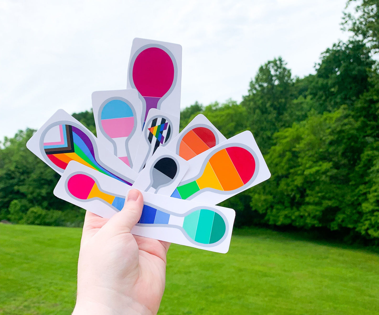 9 different pride sticker are shown on their white sticker paper being held up by a hand. The flags are pansexual, gay man, progressive pride, transgender, asexual, straight ally, rainbow, lesbian, and bisexual.