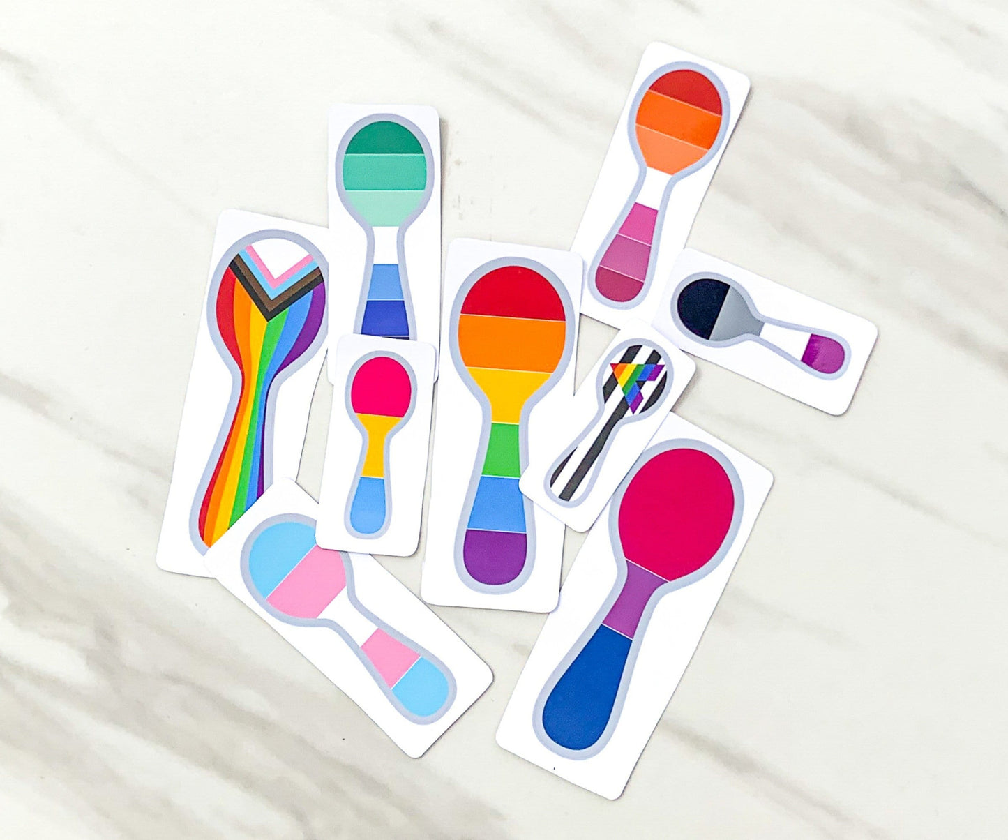 9 Stickers are shown on a marble surface. Each sticker is shaped like a spoon, with a grey border. The inside of the spoons have the flags of different LGBTQ pride flags horizontally, except for the progressive pride and straight ally flags. There are 3 sizes shown- 2 1/2 inch, 3 1/2 inch, and 4 1/2 inch.