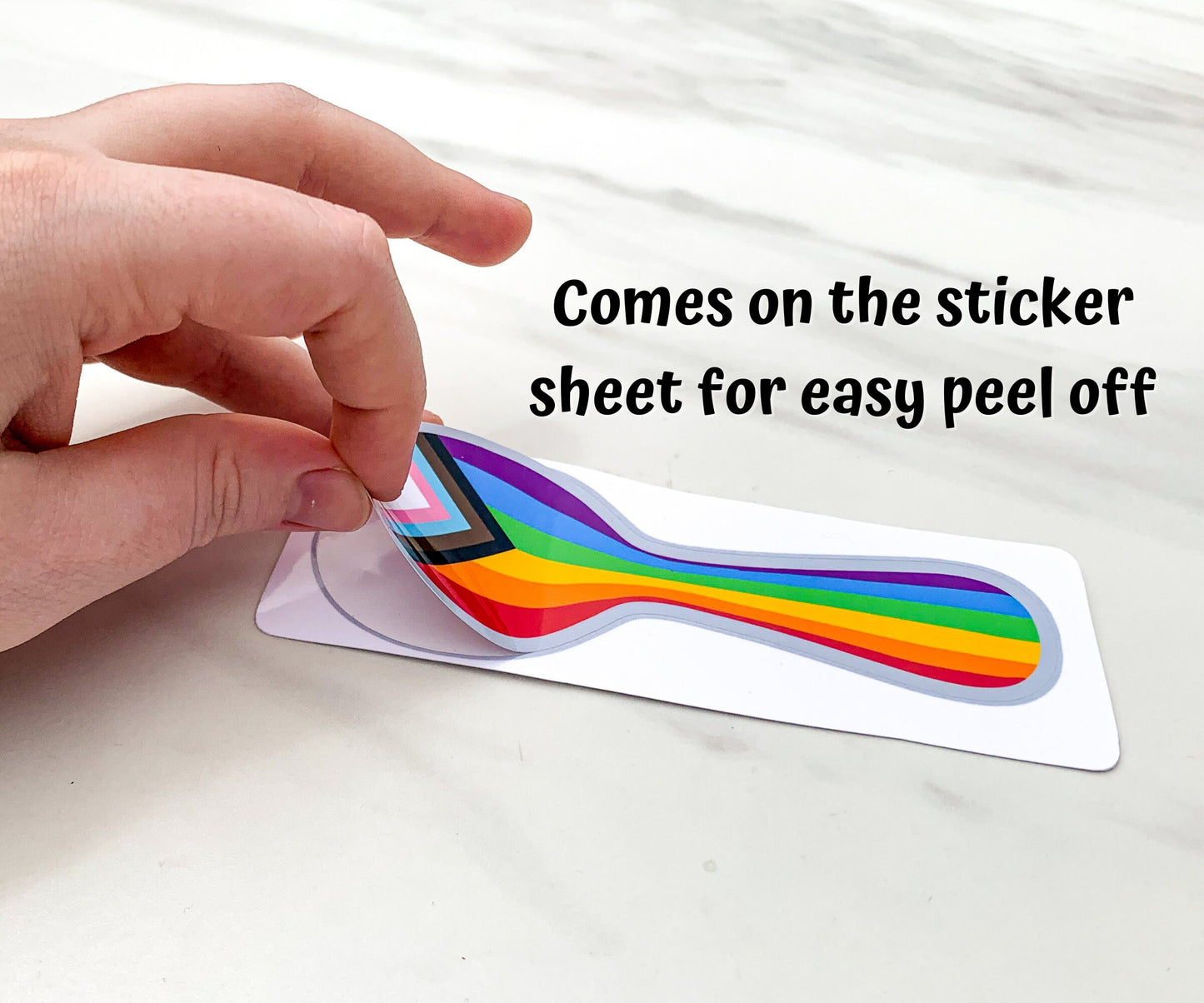 A hand is shown peeling off the progressive pride sticker from the sticker paper. A note says comes on the sticker sheet for easy peel off.