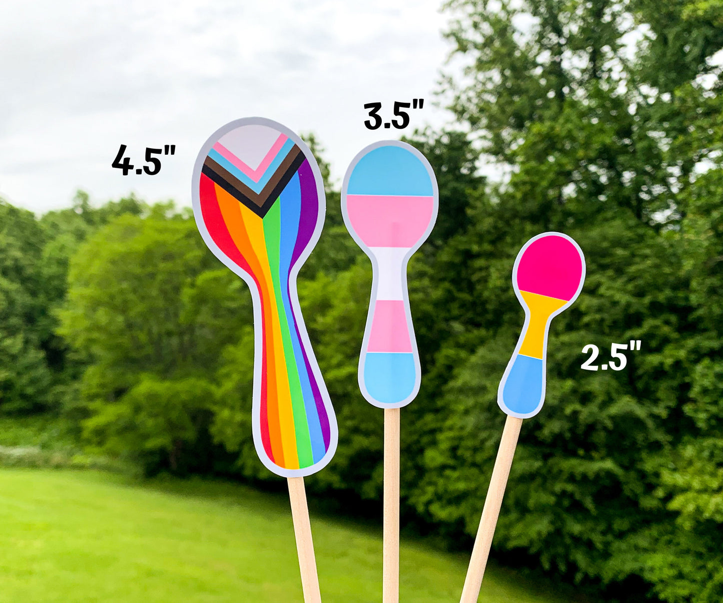 3 Pride Spoon stickers are shown on sticks. The 4.5 inch progressive pride flag- that has vertical rainbow lined, squished at the neck of the spoon. It also has triangles of the trans pride colors and brown and black at the top. The 3.5 inch transgender pride spoon has blue pink white pink blue stripes horizontally. The 2.5 inch pansexual pride spoon has hot pink yellow and blue stripes horizontally.