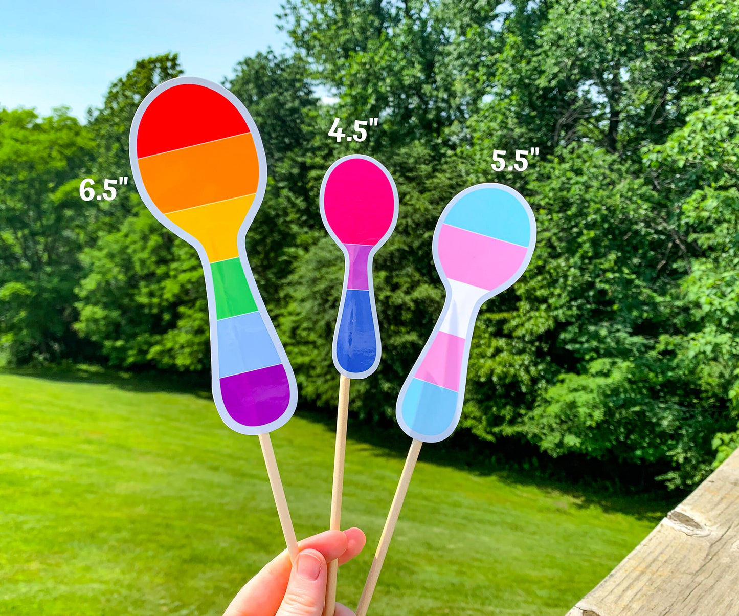 3 more Pride Spoon stickers are shown on sticks. The 6.5 inch rainbow spoon has the normal 6 rainbow colors horizontally. The 4.5 inch bisexual pride spoon has magenta, purple, and royal blue stripes horizontally with the purple being smaller. The 5.5 inch transgender spoon has the same colors and horizontal lines as the one in the previous photo, but is larger.
