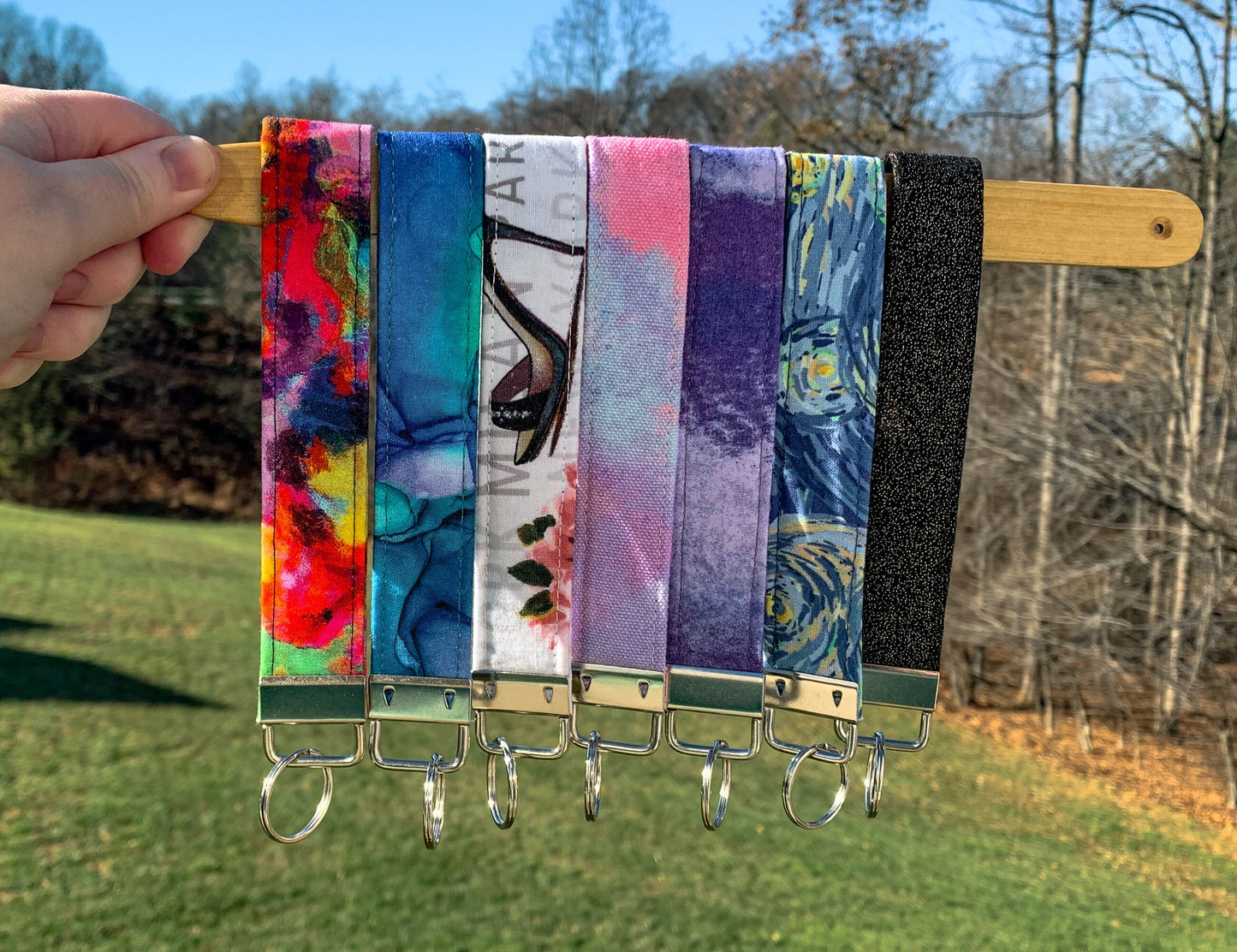 6 wristlets are shown hanging on a wood stick. The wristlets are 1 inche wide fabric folded over with a silver colored metal bracket at the bottom with a split key ring on it. The wristlets are in the colors Brushstrokes rainbow, glamour, pink clouds, brushstrokes purple, starry night,, and 2 unlisted.