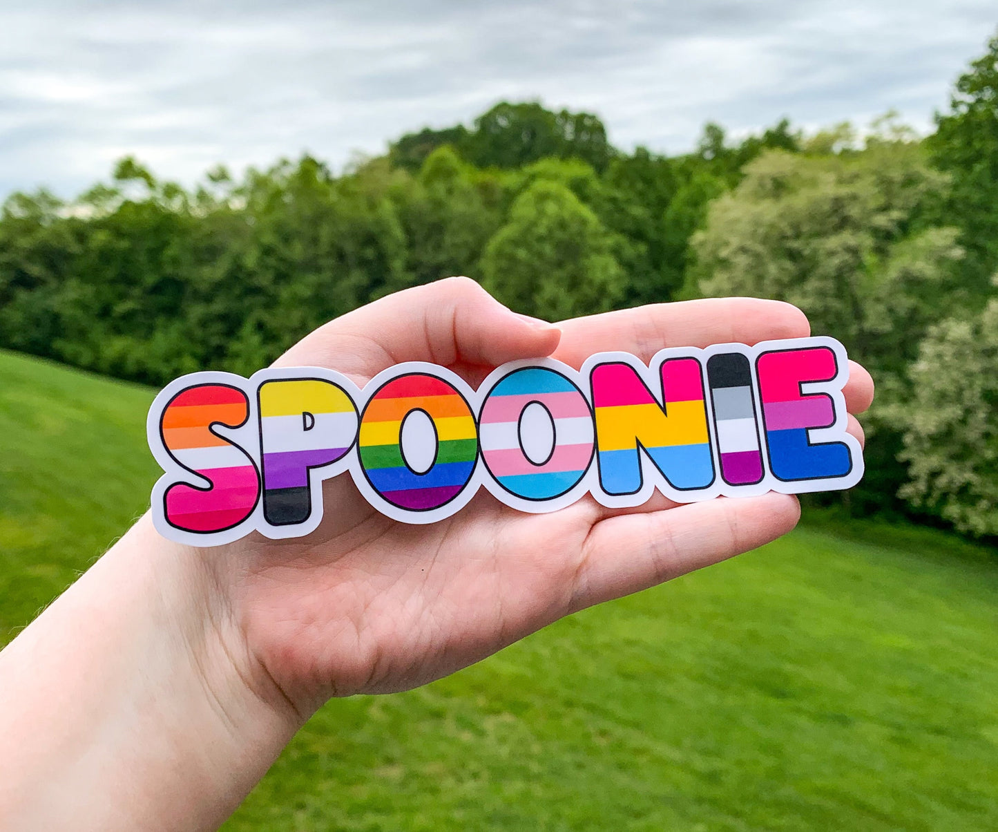 The spoonie sticker is shown being held up again, this time a hand is acting as a size comparison and backgroumd, you see trees and grass in the background.