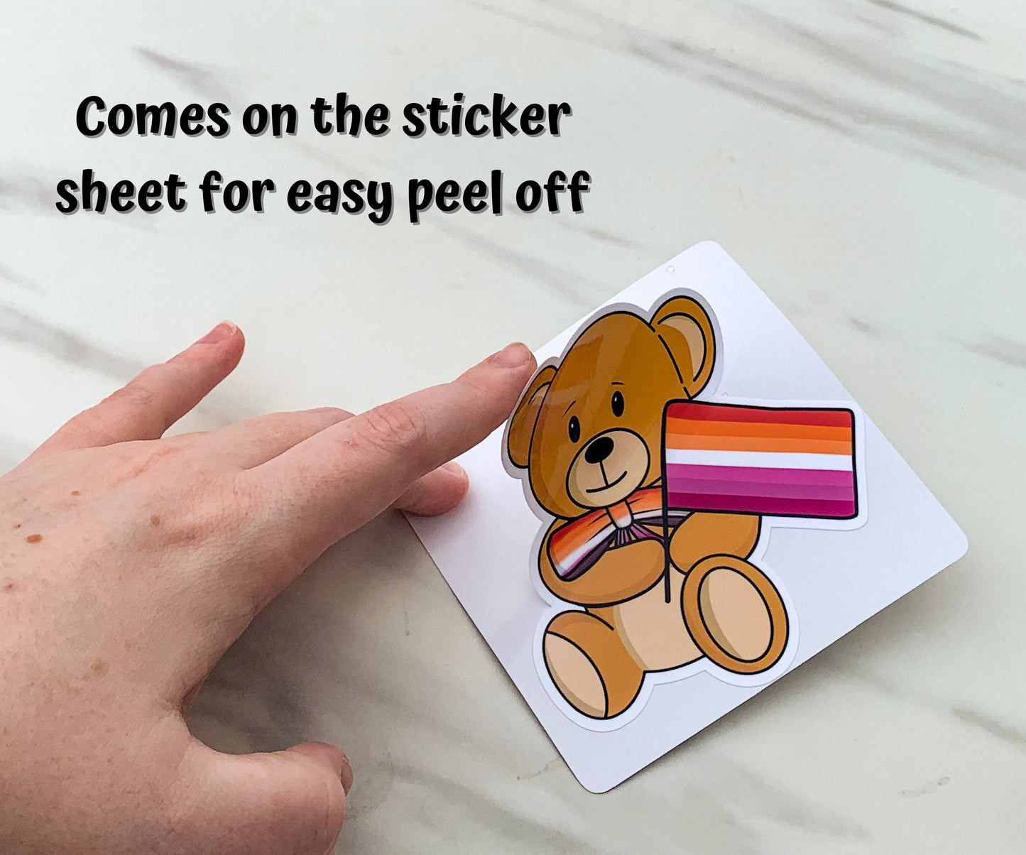 A bear holding a lesbian pride flag with a matching bow tie is shown being peeled off sticker paper. The sticker comes on the sticker sheet for easy peel off.