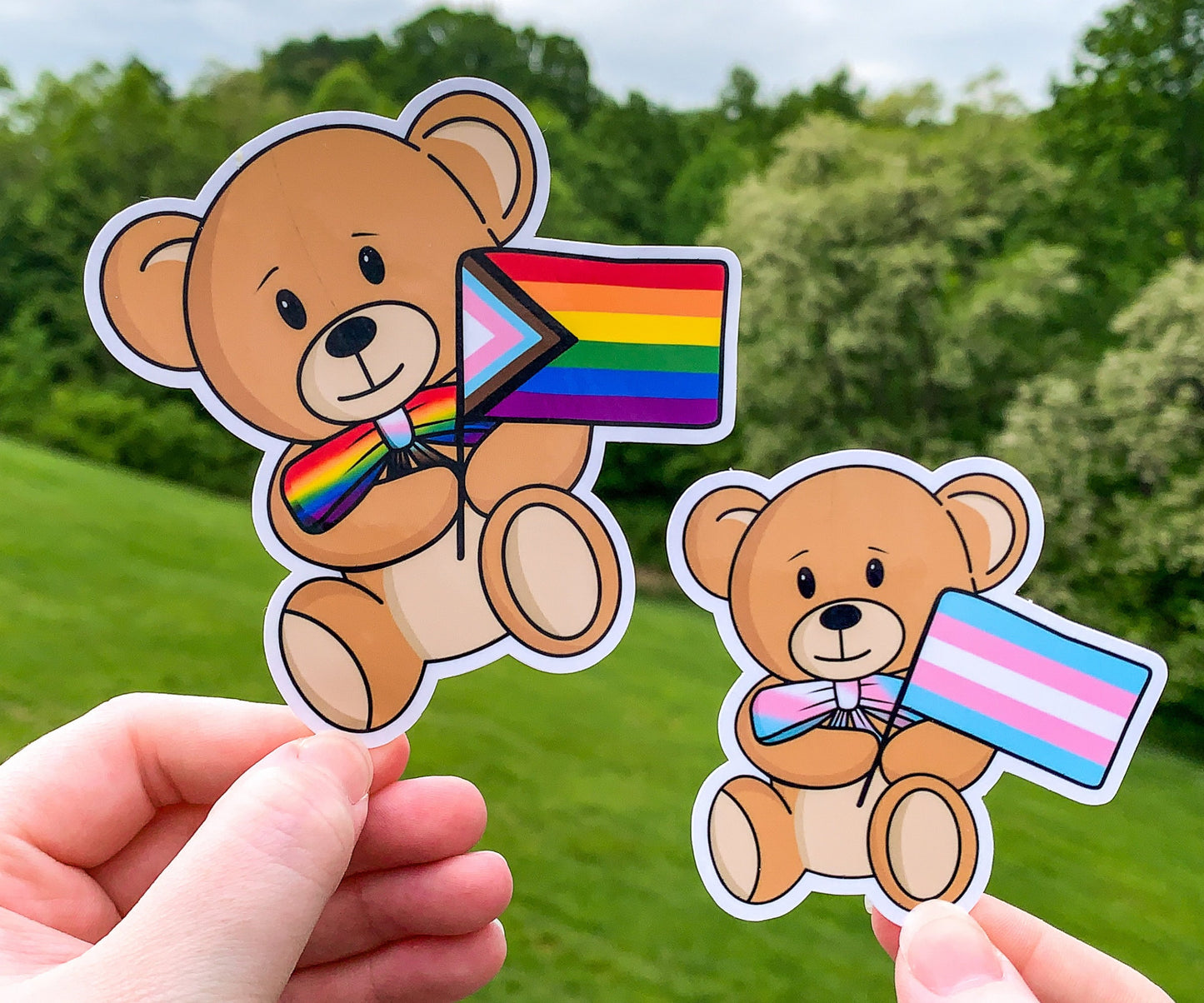 2 stickers are shown being held up. The stickers are brown teddy bears holding pride flags with a matching colored bow tie. They are very cute. The stickers have a white outline of the teddy bear.