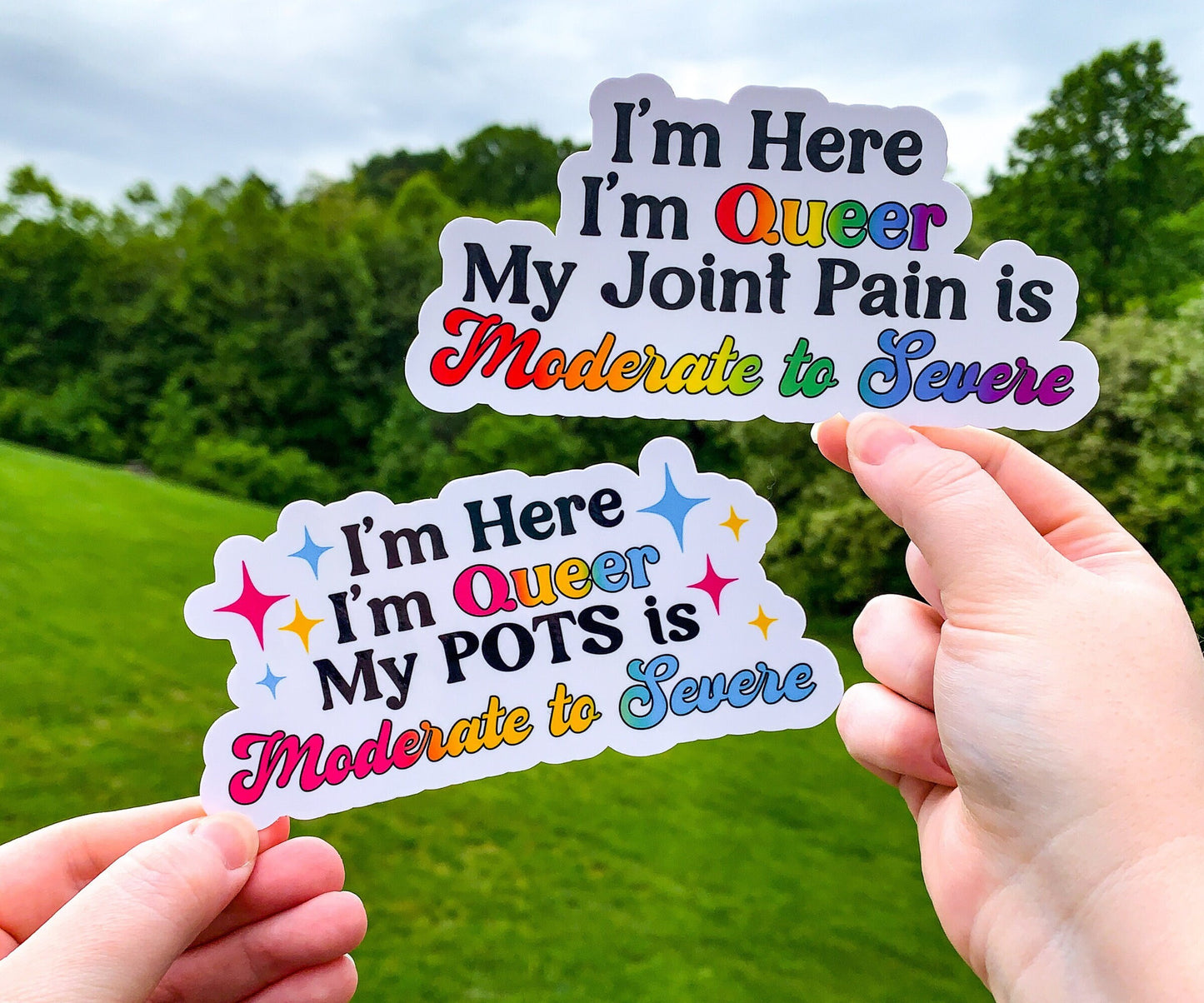 2 Stickers are shown being held up. Both have the same prompt of I'm Here I'm Queer My Blank is Moderate to Severe. Queer and Moderate to Severe have a gradient of pride flag colors. The top right one says Joint Pain in the blank part and has the rainbow pride flag. The bottom left says POTS in the blank part and has the pansexual flag colors, there are 4 sparkles in corresponding colors on each side of the text.