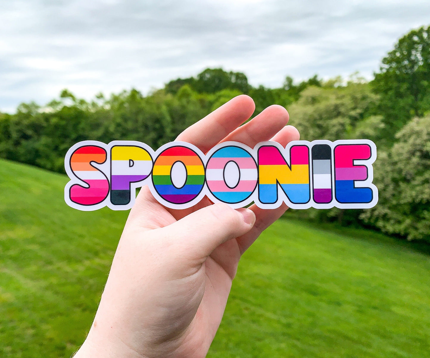 A sticker in the word spoonie is shown being held up. Each letter of Spoonie has a different pride flag. The flags in order are Lesbian, Non Binary, Rainbow, Transgender, Pansexual, Asexual, and Bisexual.
