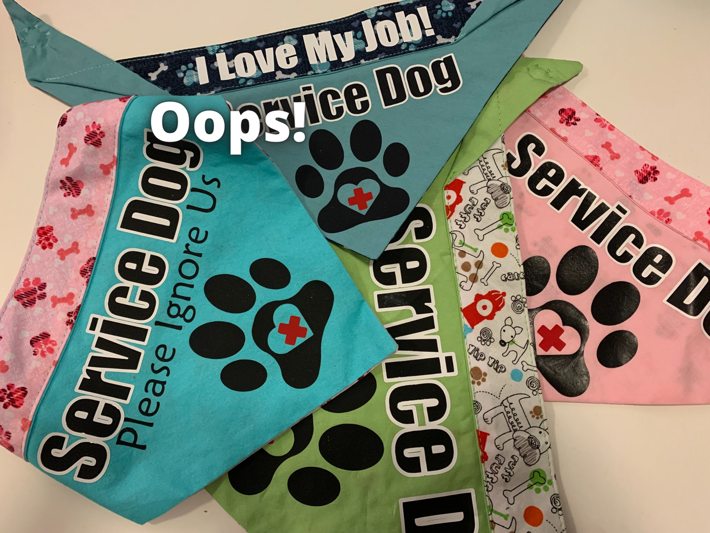 4 Bandanas are shown. One in Aqua and Pink, one in Blue and navy, another in green and white pattern, and the last at the bottom in pink. All say service dog on them with a paw, one says Please Ignore Us in addition. White letters say Oops!