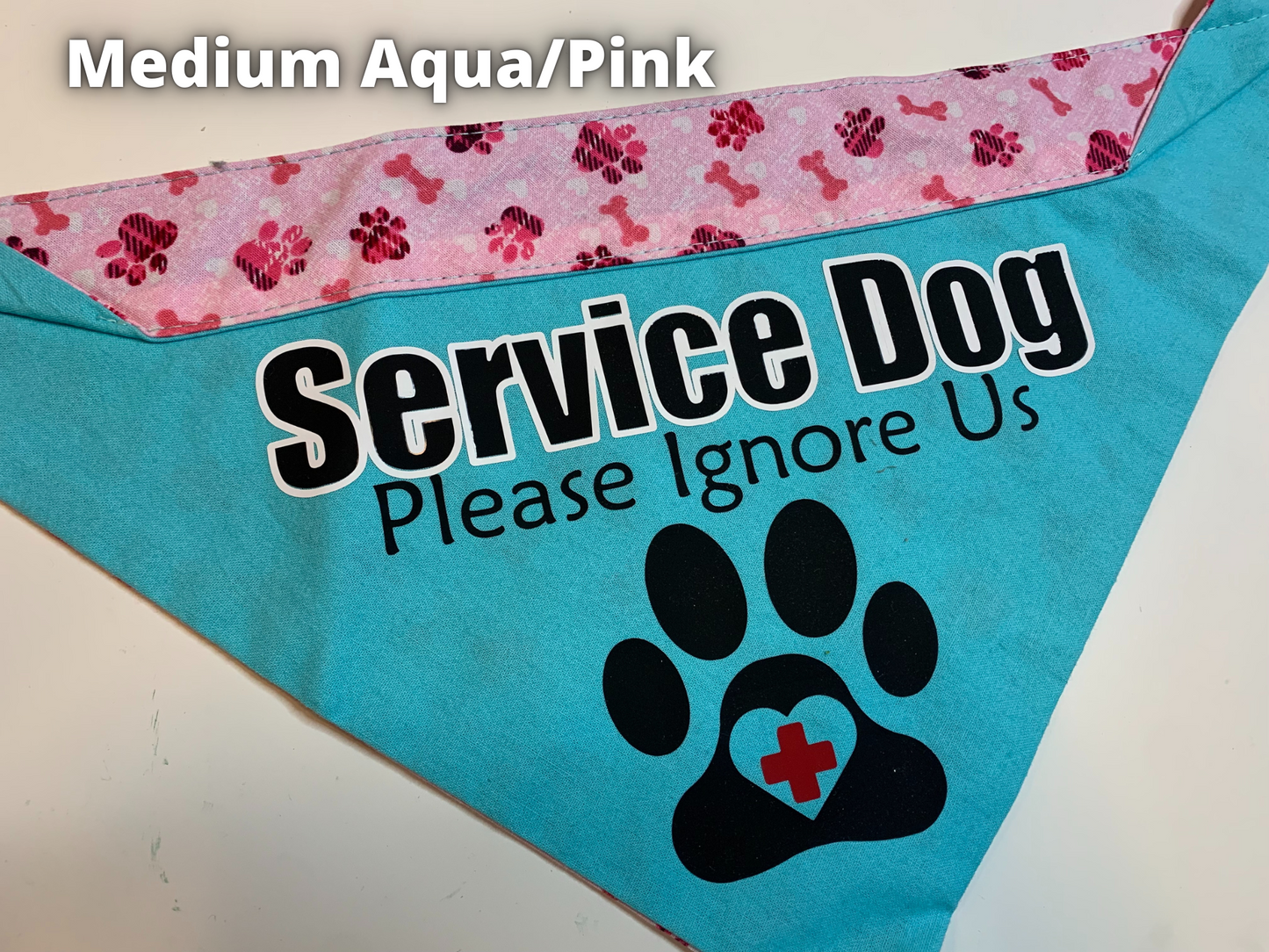 A Bandana is shown on a white surface, a label says Medium Aqua Pink. The main part of this bandana is a bright aqua color and the contrast at the top is pink paws and bones. Black text with a white outline says service dog, smaller black text below says Please Ignore Us. A black paw print below has a heart cutout in the center of the pad with a red plus in the center
