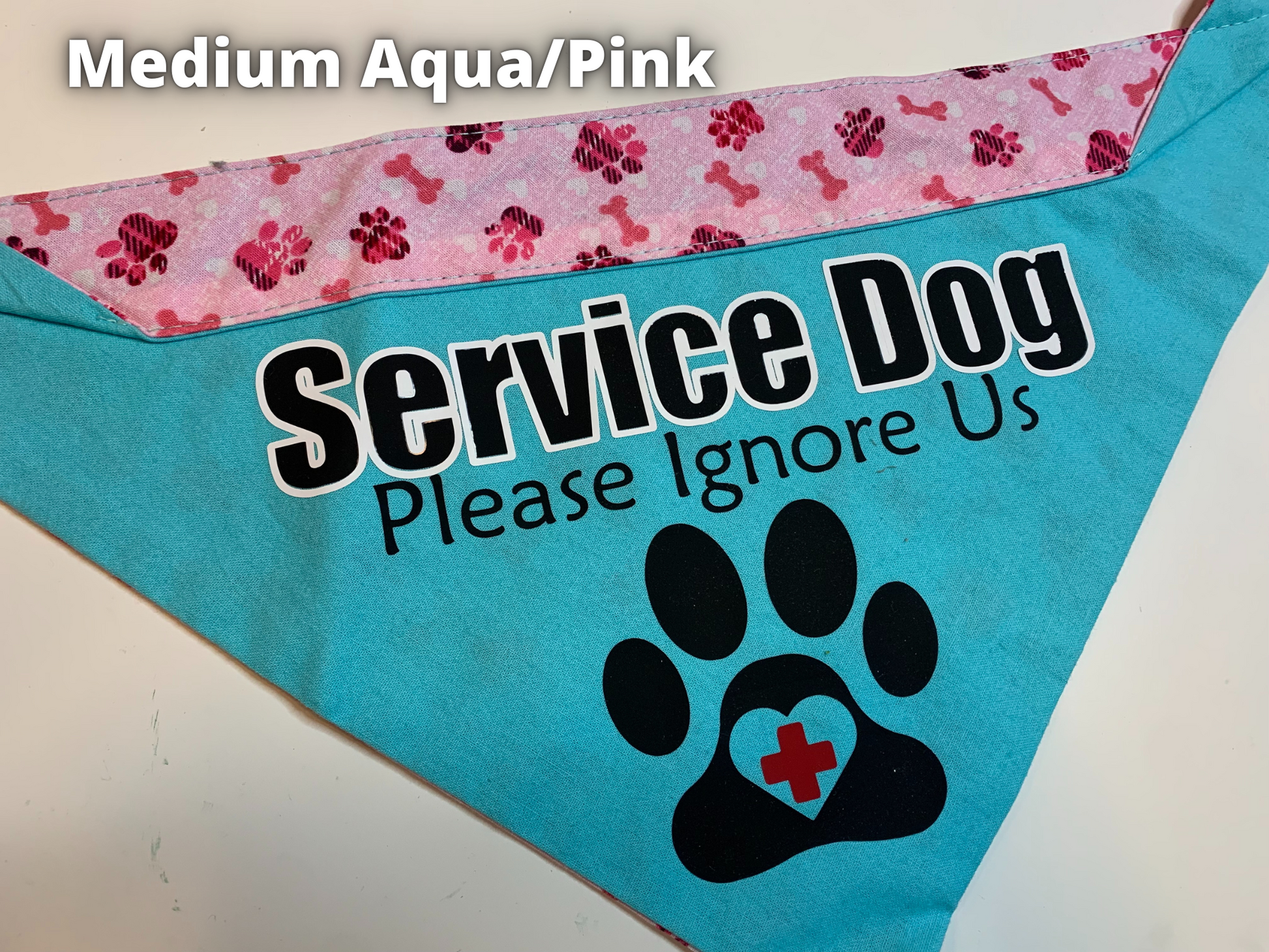 A Bandana is shown on a white surface, a label says Medium Aqua Pink. The main part of this bandana is a bright aqua color and the contrast at the top is pink paws and bones. Black text with a white outline says service dog, smaller black text below says Please Ignore Us. A black paw print below has a heart cutout in the center of the pad with a red plus in the center