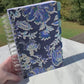 A video shows a blue and gold floral covered spiral bound book being held up, it has light and dark blue tabs that say month & trackers, then 6 months in order, and notes. An interior page is shown with days of the week, dates, what i did today, body outlines, what i ate, meds and symptom trackers and more. A grid page is shown. The top one has high medium low and is grid like, the bottom has one area per day of a month to check off. A calendar page is shown.