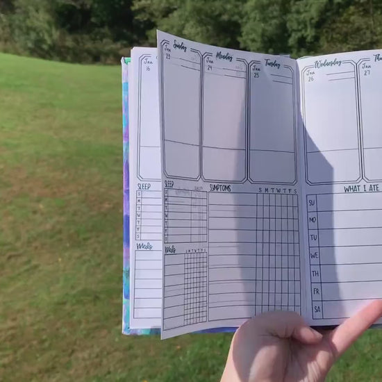 A video shows move left to right showing the main page of the symptom journal. The book is then opening laying down and is flicked to different pages to show what is in the book. The pages shown in order are the month page, main tracking page,then in the back medication and test history, doctor information, and lined pages