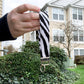 The zebra key fob is being shown held up in front of a building. It has black frayed stripes on a white background. The keychain is a loop of this fabric. It is held together at the bottom by a silver clasp with a bracket that hold a single key ring.