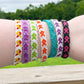 7 Bracelets are shown on a wrist. They are friendship style bracelets of knotted cotton with an awareness ribbon design. 4 of the bracelets have a white background and the ribbon colors and lime green, orange, purple, and a yellow red combo. One bracelet has a purple ribbon with a matching lighter purple background. Another has an teal ribbon with darker matching background. The last one has a black background and pink ribbon. 