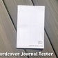 The hardcover tester is shown closed on grey wood planks. You can see the spoon culture logo on the bottom right and text that says symptom journal tester. On the left edge of the booklet you can see the back side of 3 staples. Text on the page says Hardcover Journal Tester.
