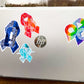 A silver laptop is shown with a 4 1/2 inch word cloud MECFS ribbon compared to a 3 1/2 inch watercolor CRPS and word cloud autism ribbon and a 2 1/2 inch word cloud POTS sticker.