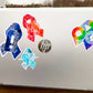 A silver laptop is shown with a 4 1/2 inch word cloud MECFS ribbon compared to a 3 1/2 inch watercolor CRPS and word cloud autism sticker, and a 2 1/2 inch word cloud POTS sticker.