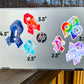 A silver laptop is shown with several stickers for size comparisons. There are 2 1/2 inch Adenomyosis and POTS stickers, 3 1/2 inch CRPS and Fibromyalgia stickers, and 4 1/2 inch Neurodiversity and ME/CFS Stickers