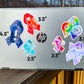 A silver laptop is shown with several stickers for size comparisons. There are 2 1/2 inch Adenomyosis and POTS stickers, 3 1/2 inch CRPS and Fibromyalgia stickers, and 4 1/2 inch Neurodiversity and ME/CFS Stickers