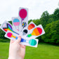 9 different pride sticker are shown on their white sticker paper being held up by a hand. The flags are pansexual, gay man, progressive pride, transgender, asexual, straight ally, rainbow, lesbian, and bisexual.