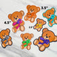 Several bears are shown with sizes. A tiny bear with a yellow ribbon is shown as 2.5 inches. 2 bears with turquoise and orange ribbons (matching outlines) are shown as 3.5 inches. The purple pink polka dot ribbon bear shown before is marked as 4.5 inches. Purple, green, and zebra ribbon bears are also shown. The zebra bear has a tighter zebra design on the bow tie versus the ribbon.