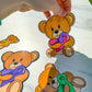 A hand is peeling up a teddy bear sticker from a marble surface. The bear has a mixed ribbon with one purple side and one pink polka dot side. The bow tie has the same dot pattern and same colors. Purple, green, and yellow ribbon bears are on the marble surface. This is to demostrate that the stickers can peel off.