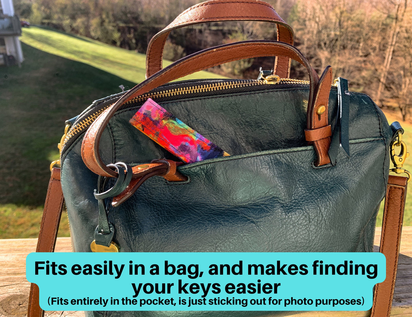 A brushstrokes rainbow wristlet is shown sticking out of the pocket of a dark teal purse. A note says fits easily in a bag, and makes finding your keys easier. Fits entirely in the pocket, is just sticking out for photo purposes.