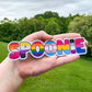 The spoonie sticker is shown being held up again, this time a hand is acting as a size comparison and backgroumd, you see trees and grass in the background.