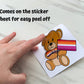 A bear holding a lesbian pride flag with a matching bow tie is shown being peeled off sticker paper. The sticker comes on the sticker sheet for easy peel off.