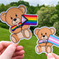 2 stickers are shown being held up. The stickers are brown teddy bears holding pride flags with a matching colored bow tie. They are very cute. The stickers have a white outline of the teddy bear.