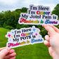 2 Stickers are shown being held up. Both have the same prompt of I'm Here I'm Queer My Blank is Moderate to Severe. Queer and Moderate to Severe have a gradient of pride flag colors. The top right one says Joint Pain in the blank part and has the rainbow pride flag. The bottom left says POTS in the blank part and has the pansexual flag colors, there are 4 sparkles in corresponding colors on each side of the text.