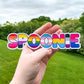 A sticker in the word spoonie is shown being held up. Each letter of Spoonie has a different pride flag. The flags in order are Lesbian, Non Binary, Rainbow, Transgender, Pansexual, Asexual, and Bisexual.